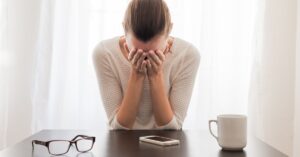 Stressed woman sitting at a table, holding her head in her hands, with glasses, a smartphone, and a white coffee mug on the table in front of her.