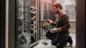 A technician kneeling beside a server rack, managing network cables with a laptop and phone—illustrating reliable, hands-on web hosting support.