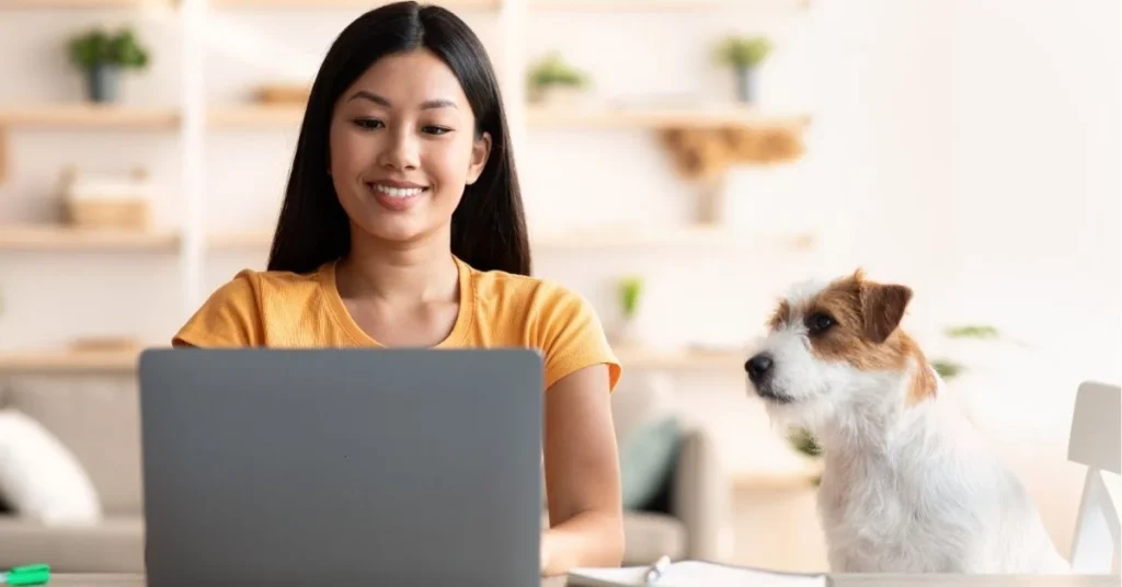 A person smiling while working on a laptop at a table, with a small dog sitting beside them, inside a cozy room.