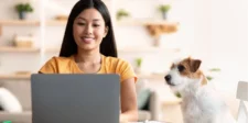 A person smiling while working on a laptop at a table, with a small dog sitting beside them, inside a cozy room.