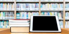 A stack of books next to a tablet on a wooden table, with blurred bookshelves in the background.