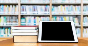 A stack of books next to a tablet on a wooden table, with blurred bookshelves in the background.