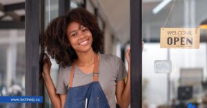 A smiling young woman with curly hair and an apron is standing in the doorway of her small business with an 'Open' sign hanging beside her—representing starting up a business.