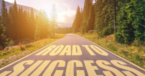 Sunny road through a forest with the words "ROAD TO SUCCESS" painted in large letters on the asphalt.