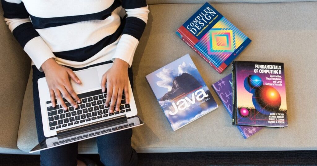 "Person using a laptop, surrounded by books on Java, compiler design, and computing fundamentals."