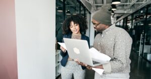 Two colleagues discussing work, with one holding a laptop and the other reviewing documents in a modern office setting.