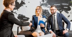 A couple meeting with a travel agent, holding passports and tickets, with a world map on the wall behind them.