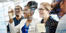 Group of four colleagues smiling and collaborating, writing ideas on a glass wall with sticky notes and markers.