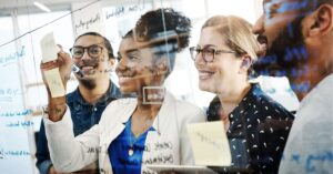 Group of four colleagues smiling and collaborating, writing ideas on a glass wall with sticky notes and markers.