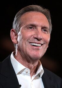 Howard Schultz, former CEO of Starbucks, smiling while speaking at an event, dressed in a black suit with a white shirt.