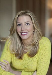 A portrait of Sara Blakely, founder of Spanx, wearing a bright yellow sweater and smiling confidently in a warmly lit indoor setting.