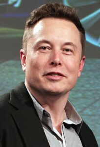 A close-up portrait of Elon Musk wearing a black blazer and a light-colored shirt, speaking at an event with a futuristic background.