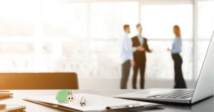 An office desk with a laptop, documents, and a cartoon bed bug illustration, symbolizing the issue of bed bugs in the workplace, while employees converse in the background.