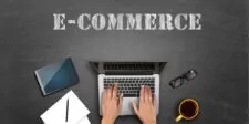 Hands typing on a laptop with "E-COMMERCE" text above. Tablet, notepad, glasses, and coffee nearby.