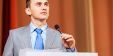 Person in a suit speaking at a podium with a microphone.