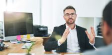 Business professional in a meeting, gesturing while speaking, with a computer in the background.