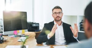 Business professional in a meeting, gesturing while speaking, with a computer in the background.