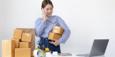 Woman on the phone holding packages, with shipping supplies and a laptop on the table.