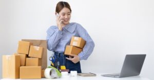 Woman on the phone holding packages, with shipping supplies and a laptop on the table.