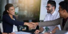 Colleagues shaking hands in a meeting room, smiling, with a whiteboard in the background.