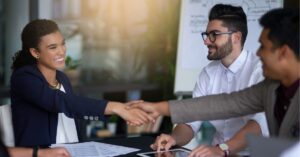 Colleagues shaking hands in a meeting room, smiling, with a whiteboard in the background.