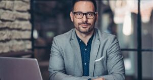 Smiling professional man in glasses and a suit, sitting at a desk with a laptop.