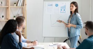 Woman presenting data on a flip chart to a group in a meeting room.