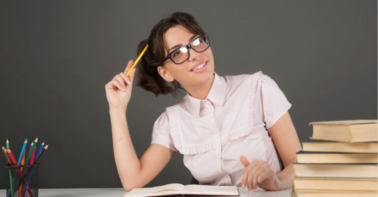 Woman wearing glasses, smiling and holding a pencil, surrounded by books and colored pencils.