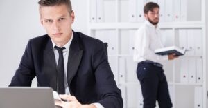 Man in a suit at a desk with a colleague in the background holding a book.