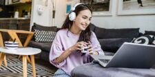 How Internet Speed Quality Impacts Hosting in Canada Young woman sitting on the floor in a cozy living room, wearing headphones and smiling while using a laptop. A cup is in her hand, and a bowl of cereal is on a small table nearby—illustrating the impact of internet speed on remote work and daily online activities.