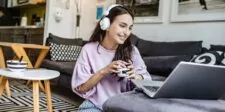 How Internet Speed Quality Impacts Hosting in Canada Young woman sitting on the floor in a cozy living room, wearing headphones and smiling while using a laptop. A cup is in her hand, and a bowl of cereal is on a small table nearby—illustrating the impact of internet speed on remote work and daily online activities.