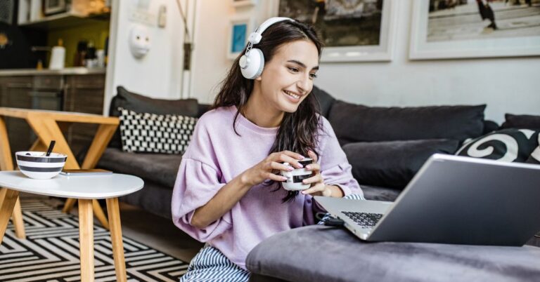 Young woman sitting on the floor in a cozy living room, wearing headphones and smiling while using a laptop. A cup is in her hand, and a bowl of cereal is on a small table nearby—illustrating the impact of internet speed on remote work and daily online activities.