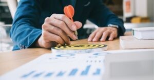 Hand holding a dart over a small target on a desk, symbolizing precision and focus.