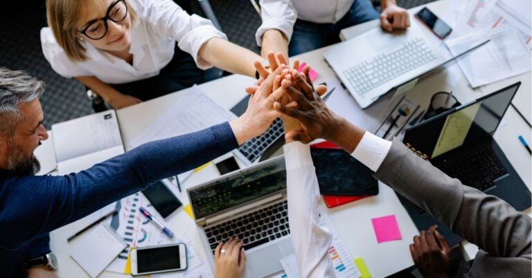 Team members joining hands over a table with laptops and documents, symbolizing teamwork.