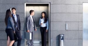 Four business professionals in suits conversing near an elevator.