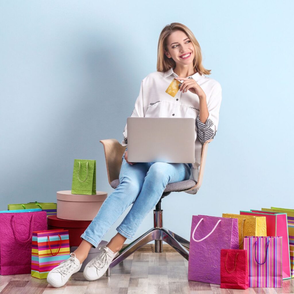 A smiling woman using a laptop while holding a credit card, surrounded by colorful shopping bags, symbolizing how copywriting services can drive online sales and boost e-commerce success.