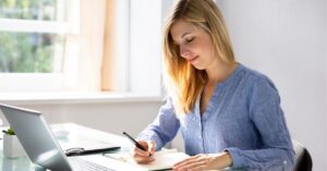 Woman writing in a notebook at a desk with a laptop.