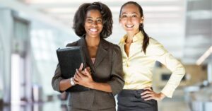 Two women smiling in an office setting. One holds a folder.