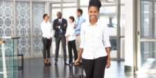 Go Big or Go Home: The Risk-Taker’s Guide to Success Professional woman confidently walking with luggage, colleagues chatting in the background.