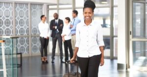 Professional woman confidently walking with luggage, colleagues chatting in the background.