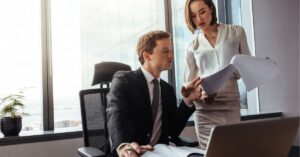Man sitting at a desk reviewing documents with a woman in an office.