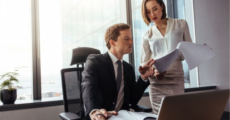 Man sitting at a desk reviewing documents with a woman in an office.