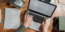 Person typing on a laptop with script pages on a wooden desk.