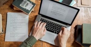 Person typing on a laptop with script pages on a wooden desk.