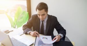 Man in a suit reviewing charts at a desk with a laptop.