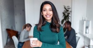 Confident young woman with long dark hair, wearing a green turtleneck, smiling while holding a coffee mug in a modern office setting. Two blurred colleagues are visible in the background.