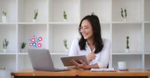 A cheerful woman is using a tablet and laptop simultaneously at a desk, surrounded by animated social media icons such as hearts and thumbs-up, symbolizing digital engagement and personalization in marketing.