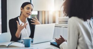 Two professionals having a discussion at a desk with a laptop.