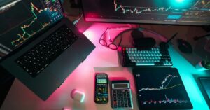 Multi-device trading setup featuring a laptop, smartphone, tablet, calculator, and monitor displaying candlestick charts and trading data in a neon-lit workspace.