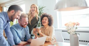 A group of colleagues laughing and collaborating around a laptop in a bright office.